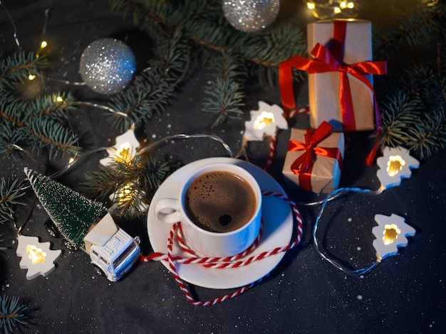 Eine weiße Tasse Kaffee, Weihnachtsgeschenke und eine weihnachtsbaumförmige Girlande auf einem dunklen Tisch am Abend. Heimelige Urlaubsatmosphäre