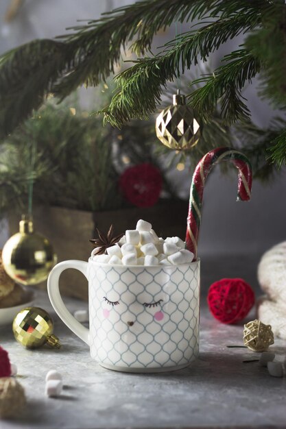 Eine weiße Tasse mit einem darauf gemalten Gesicht voller Marshmallow-Tee und einem Weihnachtsbonbonstäbchen. Rund um die Weihnachtsstimmung, Lichter, Bälle