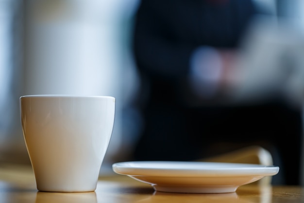 Eine weiße Tasse und Untertasse mit heißem aromatischem Kaffee steht auf einem Holztisch vor dem Hintergrund eines sitzenden Geschäftsmannes