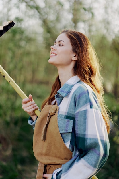 Eine wunderschön lächelnde Bäuerin in Arbeitskleidung und Schürze arbeitet draußen in der Natur und hält einen Rechen in der Hand, um Gras zu sammeln