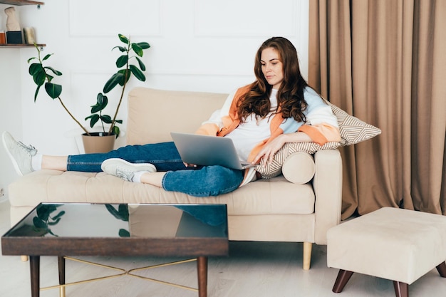 Eine wunderschöne junge Geschäftsfrau, die auf einem Sofa liegt und entfernt an einem Laptop arbeitet