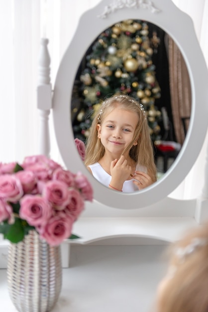 Eine wundervolle Kindheit. Nettes kleines Mädchen, das am Vorabend des Feiertags auf Weihnachten wartet. Das Kind sitzt und betrachtet sich im Spiegel und schätzt seine Schönheit.