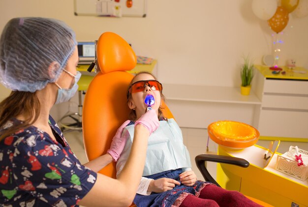 Eine Zahnärztin leuchtet eine spezielle Lampe auf die Milchzähne, um die Zähne eines kleinen Mädchens zu füllen