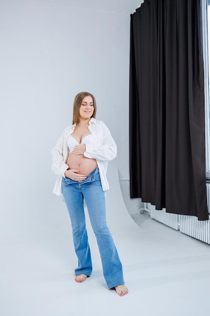 Eine zarte junge schwangere Frau in einem weißen Hemd und Jeans umarmt ihren Bauch Schwangere glückliche Frau auf weißem Hintergrund