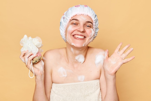 Eine zufriedene Frau, die ein Bad nimmt, einen Schwamm in der Hand hält, eine Duschhaube und ein Handtuch über dem Körper trägt, genießt hygienische Eingriffe und steht mit einem glücklichen Lächeln vor beigem Hintergrund
