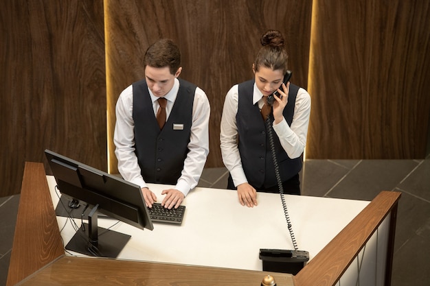 Einer von zwei jungen Hotelrezeptionisten, die an der Theke stehen und auf den Computerbildschirm schauen, während sein Kollege in der Nähe das Telefon beantwortet?
