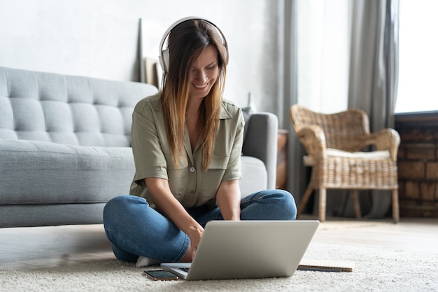 Einfach inspiriert. Überzeugte junge schöne lächelnde Frau, die am Laptop arbeitet, während sie zu Hause auf dem Boden sitzt.