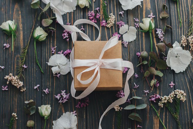 Einfache Geschenkbox mit Schleife und lila Eukalyptus-Hortensie-Rosenblättern auf dunklem Holz, flach gelegt