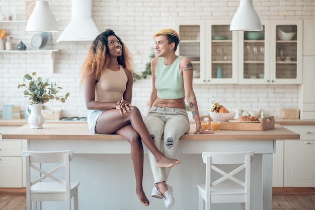 Einfache Kommunikation. Zwei junge erwachsene lächelnde Frauen in der Freizeitkleidung sitzen auf Tisch in der Küche, die angenehm plaudert