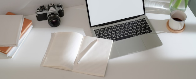 Einfacher Home-Office-Schreibtisch mit Laptop, Tagebuchheften, Kaffeetasse und Kamera auf weißem Tisch