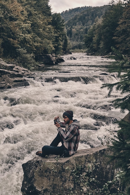 Einfangen von Erinnerungen. Hübscher junger moderner Mann, der beim Sitzen auf dem Felsen nahe dem Fluss fotografiert