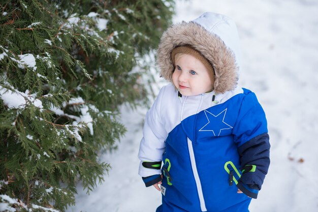 Einjähriger lachender Junge im warmen Schneeanzug, der mit weißem Schnee im Winterpark spaziert. Erster Winter und erste Kleinkinderschritte im Schnee