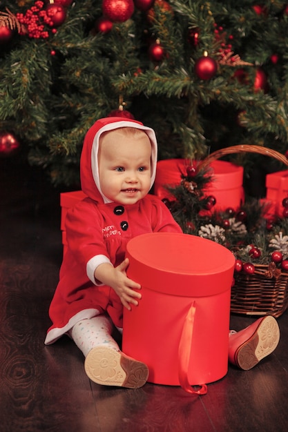 Einjähriges Kind mit blauen Augen im Weihnachtskostüm, das im Wohnzimmer mit Baum und Geschenkboxen spielt. Nettes Baby mit Emotionsfeiertagsabend. Konzept der Familienfeier von Weihnachten und Happy New Year