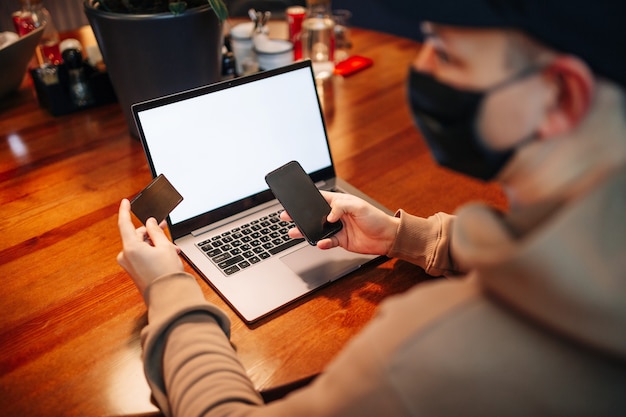 Einkaufen und Online-Zahlung mit Handy, Laptop und Kreditkarte. Mann, der Maske während der Pandemie trägt