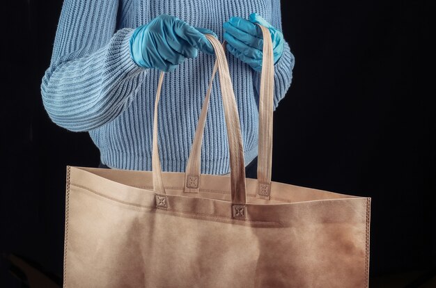 Einkaufen während der Covid-19-Pandemie. Frau in der medizinischen Maske und in den Handschuhen hält eine Tasche für das Einkaufen im Supermarkt an der schwarzen Wand.