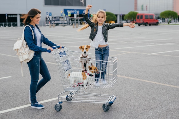 Einkaufs- und Familienkonzept. Frohe Frau trägt Trolley, in dem kleines Mädchen mit Hunden steht, posiert vor dem Hintergrund des Einkaufszentrums, freut sich über neue Einkäufe. Menschen, Glück, Ruhetag-Konzept