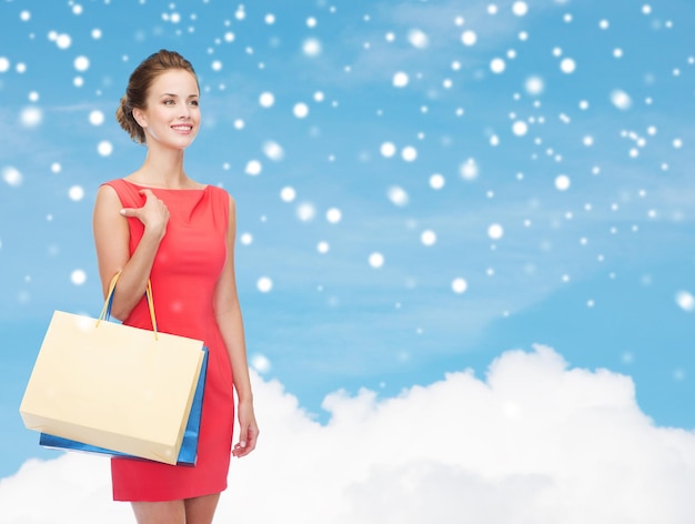 Einkaufs-, Verkaufs-, Weihnachts- und Feiertagskonzept - lächelnde elegante Frau im roten Kleid mit Einkaufstaschen über blauem Himmel mit Wolken- und Schneehintergrund