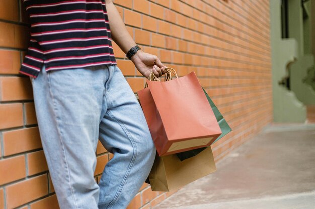 Einkaufskonzept Eine Person in hellblauen Jeans und weißen Schuhen, die sich an die Wand des Gebäudes lehnt und viele Papiereinkaufstüten hält.