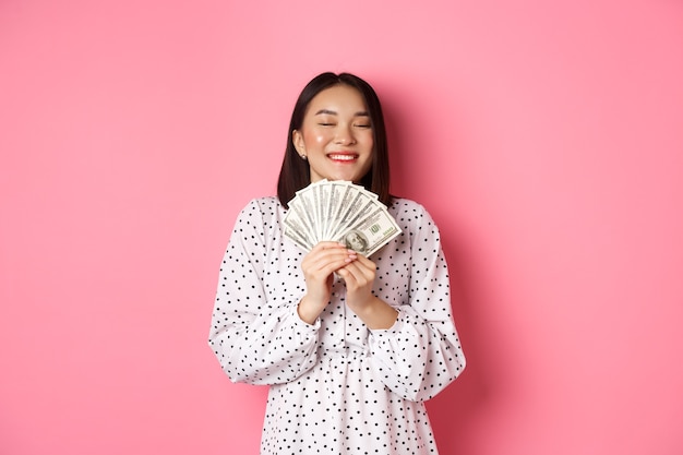 Einkaufskonzept. Glückliche und zufriedene asiatische Frau, die Preisgeld gewinnt, Dollars zeigt und sich freut, über rosa stehend.