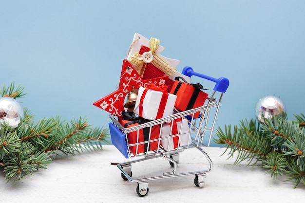 Foto einkaufswagen mit geschenkboxen und tannenzweigen mit dekoration auf blauem hintergrund. weihnachts- und neujahrsverkauf.