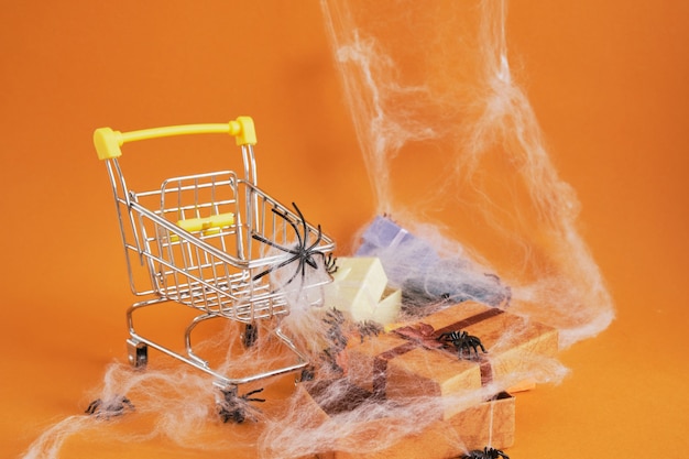 Einkaufswagen mit Spinnen- und Geschenkboxen auf braunem Hintergrund, Halloween-Shopping auf Konzept, Spinnennetz und Spinnen auf Hintergrund