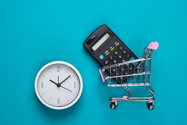 Einkaufszeit. Supermarktwagen mit Taschenrechner, Uhr auf blauer Oberfläche. Minimalismus. Draufsicht