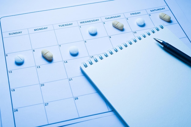 Einnahme von Pillen aufgrund des Zeitkonzepts. Nahaufnahmefoto des Kalenders mit liegenden Pillen auf den Zellen und offenem leerem Notizblock mit Schreibstift