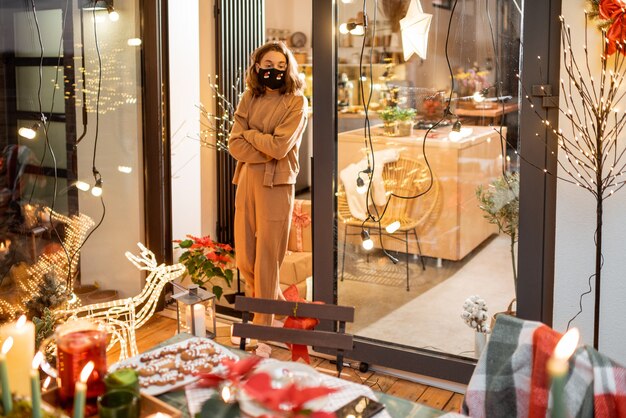Einsame Frau in Gesichtsmaske während der Neujahrsferien zu Hause auf der wunderschön dekorierten Terrasse. Konzept der Quarantäne und Selbstisolation während der Epidemie an Feiertagen