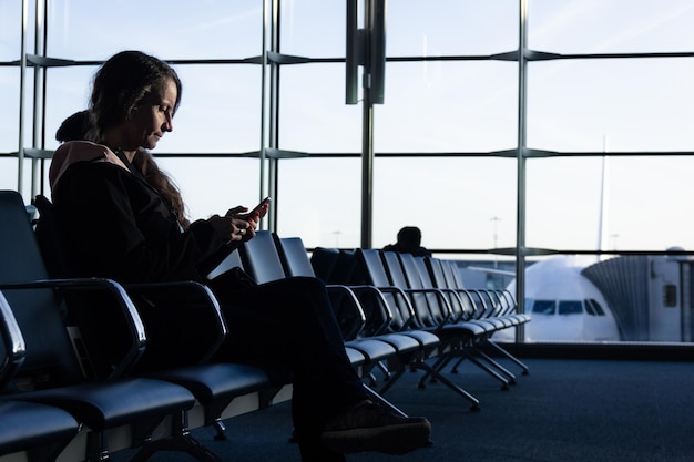 Einsame Frau sitzt am Flughafenterminal und schaut auf das Handy mit dem Flugzeug im Hintergrund