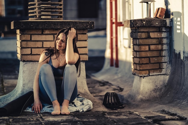 Einsame junge Frau in Depressionen sitzt auf einer Dachterrasse, verloren in schlechten pessimistischen Gedanken nach dem Yoga-Training im Freien.