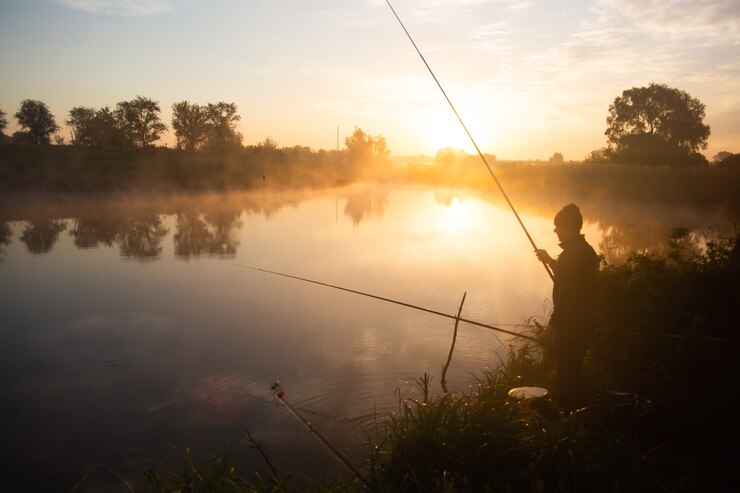 Einsamer Fischer, der am frühen Morgen kurz nach goldenem Sonnenaufgang am nebligen See angelt.