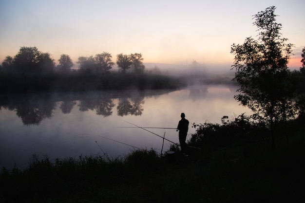 Einsamer Fischer, der am frühen Morgen kurz vor Sonnenaufgang an einem See angelt.