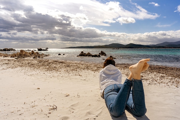 Einsames Mädchen, das auf dem Sand am Strand im Winterozeanmeer mit dramatischem blauem Himmel voller Wolken liegt.