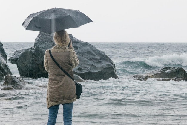 Einsamkeitskonzept für weibliche Depressionen Die Frau steht unter einem Regenschirm im Regen vor dem Hintergrund von Steinen und dem aufregenden Meer