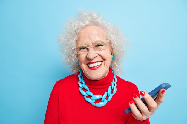 Foto einsatz moderner technologien für alle altersgruppen. reife positive grauhaarige frau mit hellem make-up in rotem pullover mit halskette verwendet smartphone wartet auf anruf lächelt positiv.