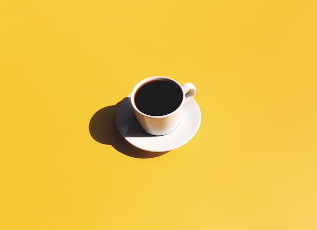 Einzelne weiße Tasse und Untertasse mit schwarzem Espresso-Kaffee aus der Draufsicht mit Schatten der Sonne auf gelb-beigem Terrakotta-Hintergrund. Foto in hoher Qualität