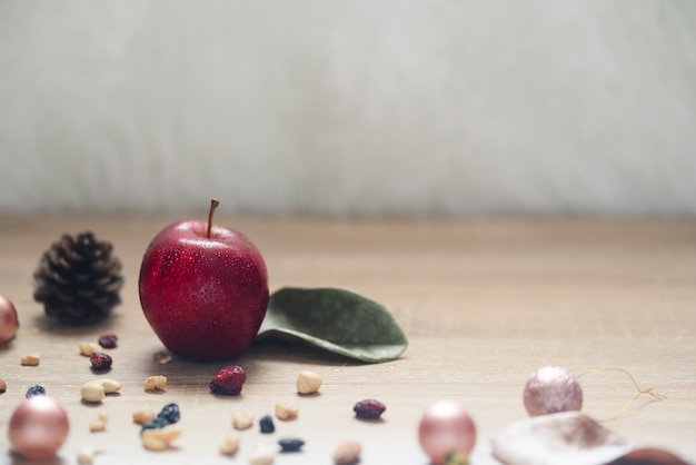 Einzelner saftiger roter Apfel mit Wasserspraytröpfchen auf Apfel einschließlich viele Mischnüsse und Rosinen, Weihnachtsbälle und Kiefernkegel auf Holztisch mit Kopienraum