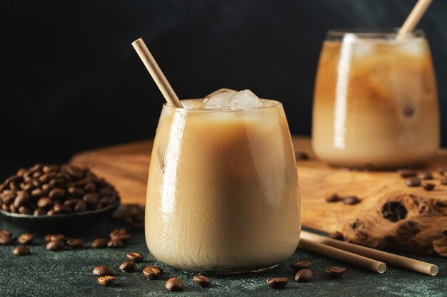 Eiskaffee in einem hohen Glas mit Sahne, über Eiswürfeln und Bohnen auf einem dunklen Betontisch gegossen, kaltes Sommergetränk mit Röhren auf schwarzem Hintergrund mit Kopierplatz