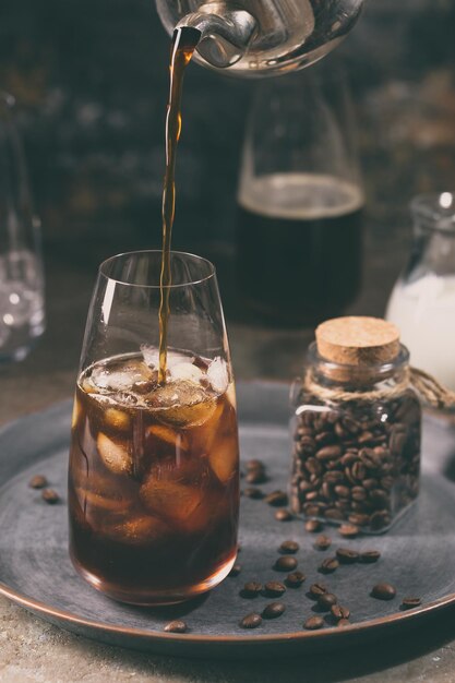 Eiskaffee kaltes Sommergetränk in einem hohen Glas mit strömendem Kaffee und Kaffeebohnen auf Steinhintergrund.