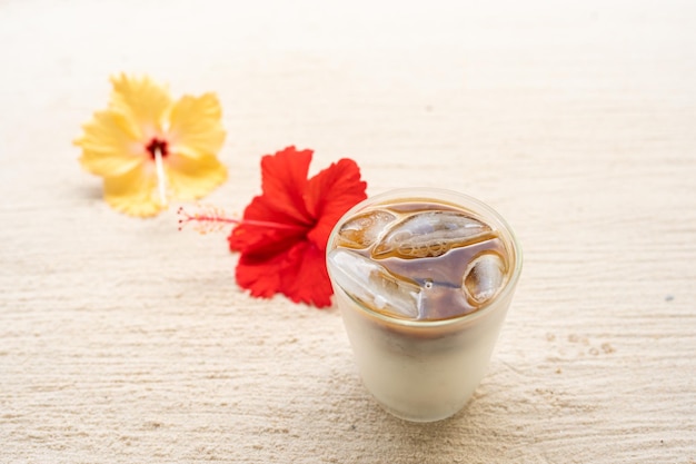 Eiskaffee mit Milch neben Hibiskus am Sandstrand.