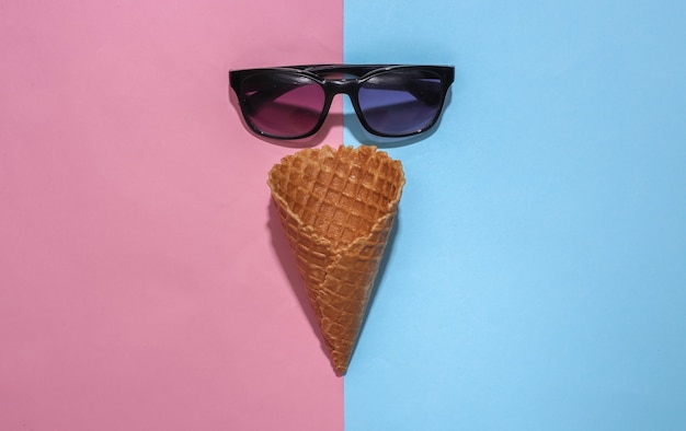 Eiswaffelkegel und Sonnenbrille auf rosa und blauem hellem Pastellhintergrund mit tiefem Schatten, Draufsicht. Flache Sommerkomposition