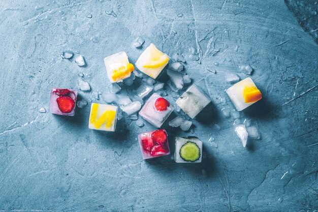 Eiswürfel mit Frucht und unterbrochenem Eis auf einer blauen Steinoberfläche. Minze, Erdbeere, Kirsche, Zitrone, Orange. flach lag, Ansicht von oben
