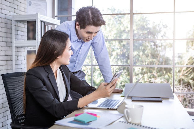 Ejecutivo de la empresa Joven empresario asiático y coaching secretaria personal asistente empresaria socios mientras trabajan juntos con mantener smartphone y estrategia en beneficio empresarial en la oficina.