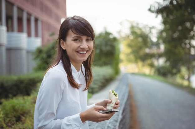 Ejecutivo sonriente con rollo de verduras mientras usa el teléfono móvil en la carretera