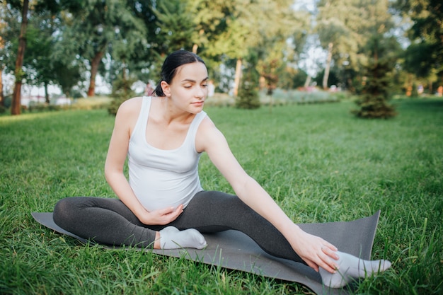 Ejercicio agradable joven de la mujer embarazada en parque afuera. Ella alcanza los pies con una mano. Otro se aferra al vientre. La modelo es tranquila y pacífica. Ella se sienta en el compañero de yoga.