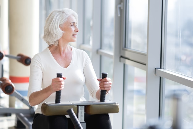 Ejercicio agradable. Mujer delgada alegre encantada mirando por la ventana y usando equipos de gimnasia mientras hace ejercicio.