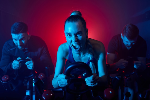 Ejercicio de cardio en bicicleta en el gimnasio de la mujer y los amigos que tienen un entrenamiento intensivo, tomando la pérdida de peso con la máquina aeróbica para el cuerpo delgado, están gritando