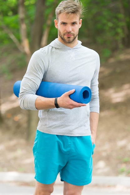 Ejercicio de estiramiento de los músculos de pilates. Deportista lleva esterilla de yoga para ejercicios al aire libre. Concepto de yoga al aire libre. La cara tranquila del atleta hombre lleva la estera para estirar el fondo de la naturaleza. Entrenador de atleta deportista.