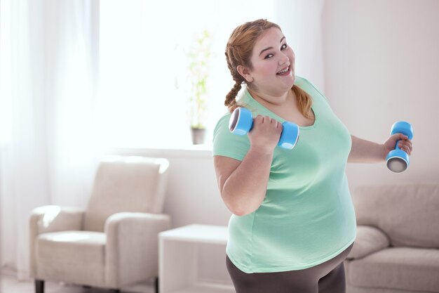 Foto ejercicio favorito. sonriente mujer pelirroja inclinada hacia la derecha mientras hace ejercicios con pesas