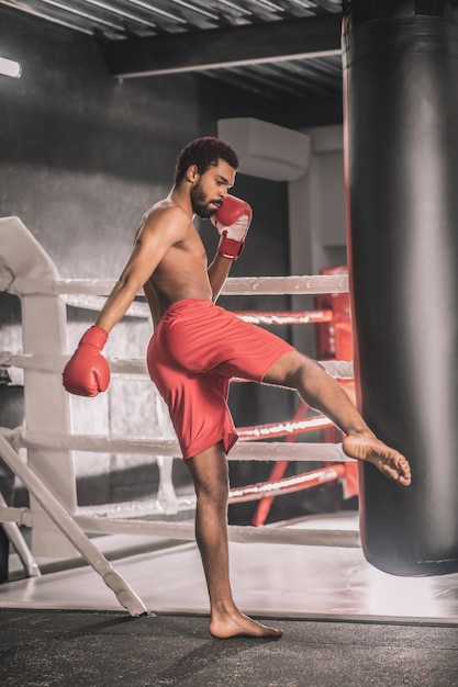 Ejercicio. Joven kickboxer de piel oscura pateando un saco de arena con la pierna